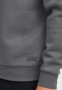 Sweat-shirt gris en tissu doux, avec un ourlet et des poignets côtelés, et une petite étiquette noire en bas.
