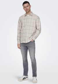 Cămașă bej cu nasturi, cu un model în grilă, combinată cu blugi gri deschis și adidași albi. Cămașa are o croială lejeră și guler.