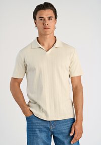 Ljusbeige polo-tröja gjord av texturerat tyg med kort krage. Har ett subtilt vertikalt mönster och är kombinerad med blåjeans.