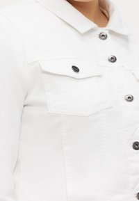 Närbild av en vit jeansjacka med metallknappar och framfickor på en persons överkropp.