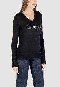 Schwarzer Pullover mit V-Ausschnitt, der das "Guess"-Logo aus Strasssteinen auf der Vorderseite trägt. Langärmlig mit geripptem Saum und Bündchen.