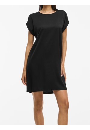 Woman wearing a loose, short-sleeve black dress with rolled sleeves, standing against a plain white background.