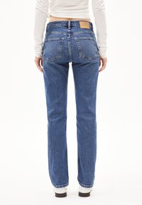 Pantalones vaqueros azules de mezclilla con corte de pierna recta, diseño de cinco bolsillos y una parche de cuero marrón claro en la parte trasera de la cintura. El material parece resistente.