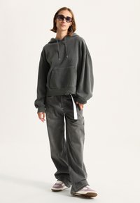 Oversized dark gray hoodie with a front pocket, paired with loose gray jeans and multicolored sneakers, featuring a white drawstring.