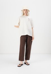 A person with long light hair stands in a relaxed pose, wearing an oversized cream t-shirt, brown pants, and a beige bucket hat.
