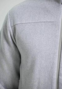 Šedá fleecová bunda s hladkým, strukturovaným povrchem a předním zipem. Má jednoduchý design a jemný ševový detail na rameni.