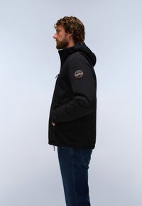 Hombre con cabello rizado y barba, vistiendo una chaqueta negra con capucha y un parche redondo en la manga y vaqueros azules, de perfil.