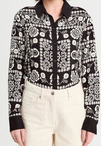 Chemise à motifs noirs et blancs avec des motifs floraux et géométriques, boutonnée à l'avant, avec un col noir. Associée à un pantalon beige taille haute.
