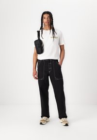 Black pants with white stitching, white t-shirt with logo, and a black crossbody bag. Sneakers feature a beige and black design.