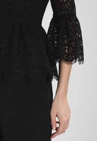 Černý krajkový top s peplum designem, propracovanými květinovými vzory, rozšířenými rukávy a třásňovým lemem.