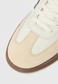 Beige-weißer Sneaker mit glattem Lederobermaterial und Wildlederakzenten, ausgestattet mit flachen weißen Schnürsenkeln und einer texturierten braunen Gummisohle.