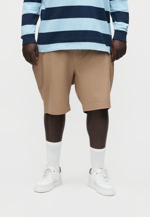 Άνδρας που φοράει μπεζ σορτς μέχρι το γόνατο, λευκά αθλητικά παπούτσια, λευκές κάλτσες και μακρυμάνικο πουκάμισο με φαρδιές οριζόντιες μπλε ρίγες.