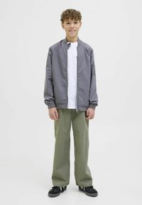 Svetlo siva bomber jakna s kapuco, nosena preko bele majice, uparena sa slobodnim zelenim pantalonama i crnim patikama. Kovrdžava kosa.