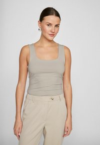 Beige fitted tank top with a square neckline. Model wears tailored beige trousers, showcasing a smooth fabric and minimalistic design.