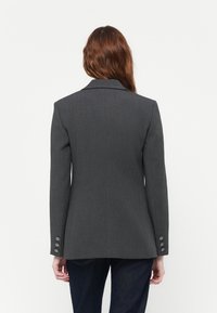 Blazer grigio con revers classico, forma sartoriale, tessuto testurizzato e tre bottoni decorativi su ciascun polsino. Vista posteriore semplice.