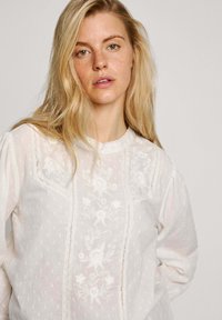 Blusa bianca ricamata con motivi floreali, tessuto testurizzato, scollatura tonda con bordi ondulati e maniche lunghe.