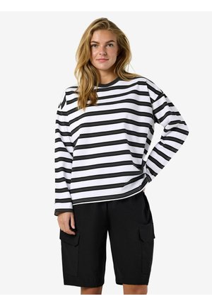 Młoda kobieta o długich blond włosach, ubrana w biało-czarną koszulkę w poziome paski z długim rękawem oraz czarne bermudy do kolan.