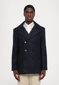 Námornícky vlnový dvojradový kabát s veľkými klopami, štyrmi čiernymi gombíkmi a bočnými vreckami. V kombinácii s košeľou so vzorom.