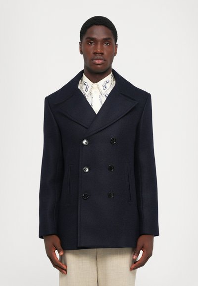 Jūras zila vilnas dubultās krūšu pakoja ar lieliem atlazēm, četriem melniem pogām un sānu kabatām. Apakšā valkāta rakstota krekls.