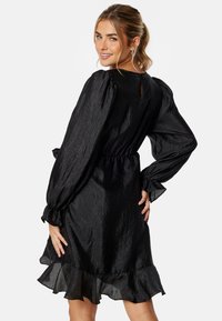 Robe noire à manches bouffantes, taille froncée et ourlet volanté. Fabriquée en tissu texturé avec un léger éclat. Détail de dos en forme de trou de serrure.