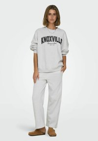 Felpa grigia chiara con testo "KNOXVILLE" in nero, abbinata a pantaloni da tuta grigi. Tessuto morbido, vestibilità comoda e dettagli con zip laterali sui pantaloni.