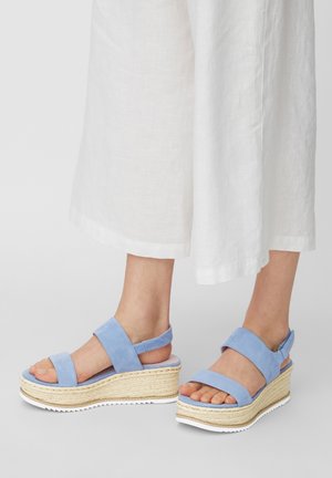 Pieds portant des sandales à plateforme bleu clair avec des semelles en jute tissé et des larges brides, associés à un pantalon blanc large et raccourci.