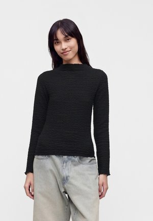 Femme portant un haut noir texturé à manches longues et un jean large délavé clair, debout devant un fond clair uni.