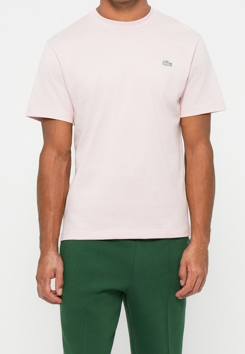 Homme portant un t-shirt rose clair à manches courtes avec un petit logo vert sur la poitrine et un pantalon vert, debout devant un fond uni.