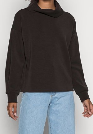 Personne portant un pull noir ample à manches longues avec un col large et un jean bleu clair taille haute sur un fond uni.