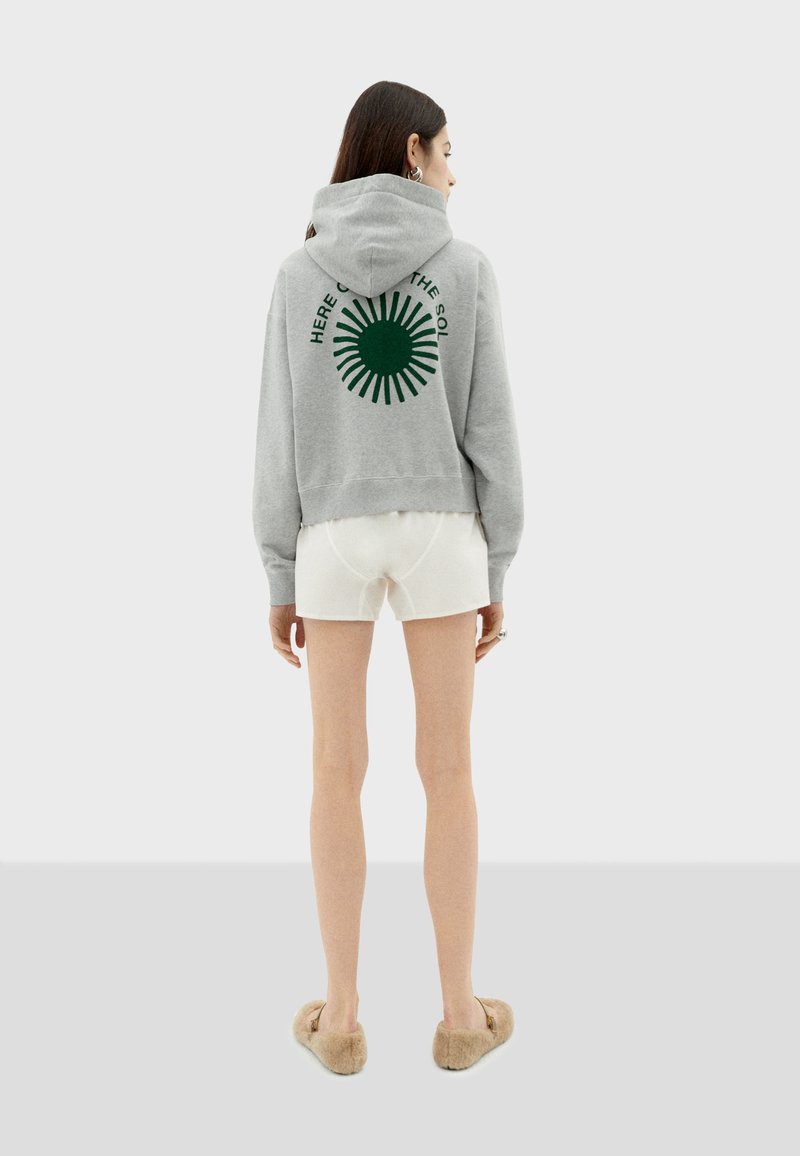 Haut à capuche gris avec un graphisme de soleil vert dans le dos, présentant une capuche à cordon de serrage, des poignets côtelés et une coupe décontractée. Associé à un short blanc.