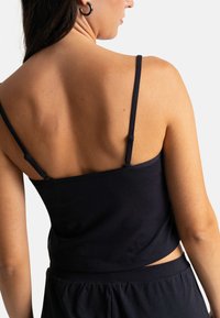 Top noir court avec fines bretelles, fabriqué en coton doux et au design sans couture. La vue de dos montre un style minimaliste avec des lignes épurées.