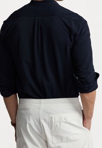 Mężczyzna w czarnej koszuli z długim rękawem, podwiniętymi rękawami oraz białych spodniach z tylną kieszenią i guzikiem, stojący z ręką w kieszeni.