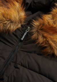 Sort pufferjakke med lynlås foran og orange-brun faux fur kant omkring hætten. Tekstureret stof med quiltet mønster.