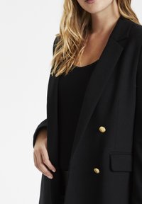 Svart kavaj med guldfärgade knappar, strukturerade slag, långa ärmar och skräddarsydd passform. Slät yta med en enda framficka.