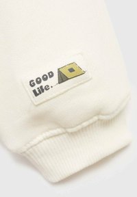 Tessuto bianco con polsino a coste presenta un piccolo etichetta che raffigura una tenda gialla e la scritta "GOOD Life". Cucita su una manica.