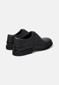 Un paio di scarpe in pelle nere con lacci, dotate di suole spesse e testurizzate, viste dal retro e di lato su uno sfondo bianco.