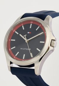 Ανδρικό ρολόι Tommy Hilfiger με μαύρο καντράν, κόκκινο εξωτερικό δακτύλιο, ασημένιους δείκτες και μπλε λουράκι σιλικόνης.