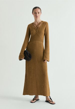 Femme portant une longue robe en maille marron aux manches évasées, tenant une pochette noire ornée de perles et portant des sandales noires à brides.