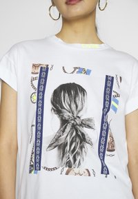 Camiseta blanca con un gráfico en blanco y negro del cabello de una mujer atado con un lazo. Acentuada con cadenas y patrones coloridos.