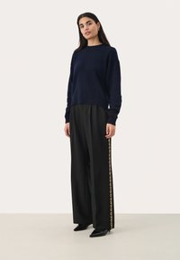 Pull en maille marine à col rond, associé à un pantalon large noir avec une bande dorée sur le côté, et des escarpins noirs à bout pointu.