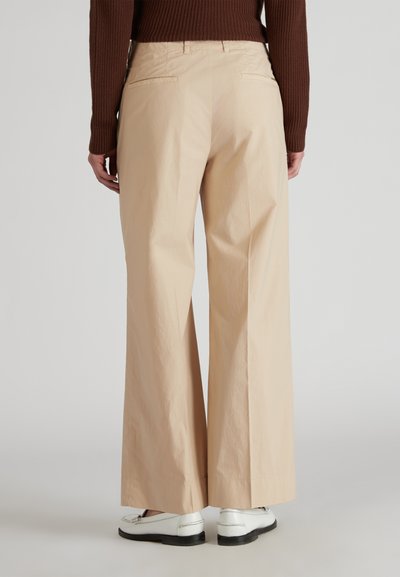 Pantalones de pierna ancha, beige, confeccionados en una tela ligera, con dos bolsillos traseros y una textura suave. Combinados con mocasines blancos.