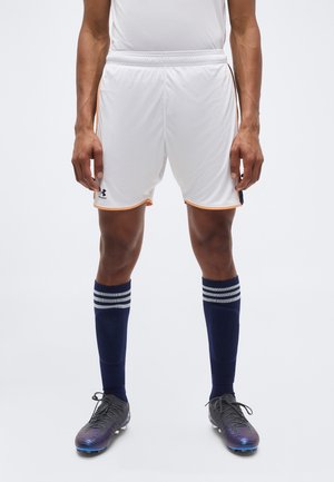 Άνδρας αθλητής που φοράει λευκά αθλητικά σορτς με πορτοκαλί περίγραμμα, ναυτικό μπλε κάλτσες μέχρι το γόνατο με λευκές ρίγες και μαύρα ποδοσφαιρικά παπούτσια.