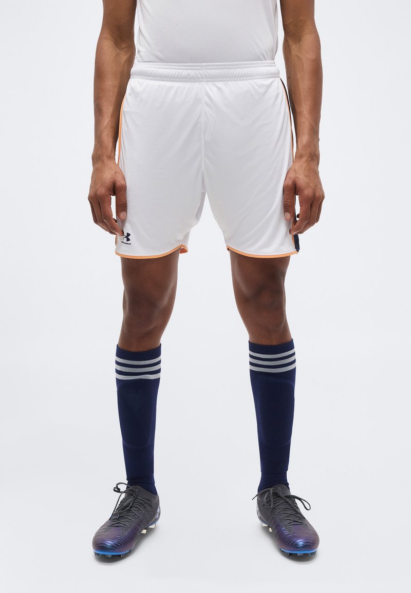 Άνδρας αθλητής που φοράει λευκά αθλητικά σορτς με πορτοκαλί περίγραμμα, ναυτικό μπλε κάλτσες μέχρι το γόνατο με λευκές ρίγες και μαύρα ποδοσφαιρικά παπούτσια.