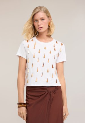 Jonge vrouw met blond haar die een wit kortemouwtop draagt met gouden druppelversieringen en een bruine rok met een geplooid detail.