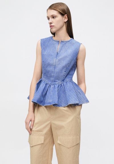 Γυναίκα που φοράει μπλε και λευκό ριγέ αμάνικο τοπ με πεπλούμ και μπεζ παντελόνι cargo, κοιτάζοντας ελαφρώς πλευρικά πάνω σε απλό φόντο.