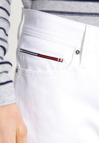 Vita denimbyxor med en liten framficka som har röd, vit och marinblå randig broderi samt metallknappdetaljer.