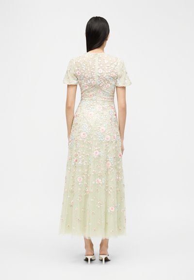 Γυναίκα με μακριά σκούρα μαλλιά φοράει διαφανές, παστέλ πράσινο φόρεμα με κεντήματα λουλουδιών, κοντά μανίκια και λευκά παπούτσια με ψηλό τακούνι, στέκεται με την πλάτη γυρισμένη.