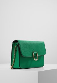 Grön texturerad läderhandväska med ett spänne i guldton, svart kant och en kedjeaxelrem. Rektangulär form med lockstängning.