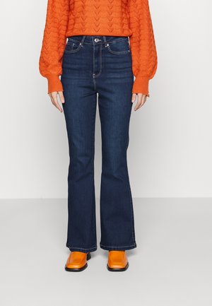 Osoba ubrana w ciemnoniebieskie, wysokie dzwony jeansowe, pomarańczowy sweter z dzianiny oraz jasnopomarańczowe buty o kwadratowym nosku, stojąca na tle jednolitej białej ściany.