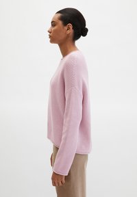 Femme aux cheveux foncés en chignon, portant un pull en tricot rose clair et un pantalon beige, debout de profil sur un fond blanc uni.